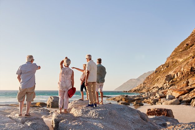 Rear View Of Senior Friends Standing On Rocks On Summer Group Vacation Looking Out To Sea Rear View Of Senior Friends Standing On Rocks On Summer Group Vacation Looking Out To Sea