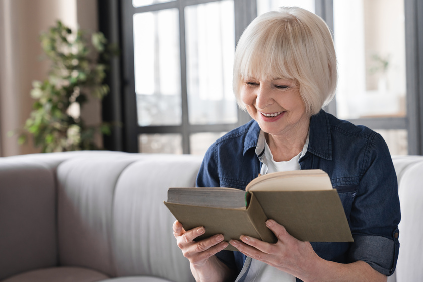 Happy old aged elderly senior woman sitting on a sofa and reading an interesting book enjoying the author’s writing style. Grandmother relaxing at home, free time. The Importance Of Onsite Library In Park Cities, TX Senior Living For Residents