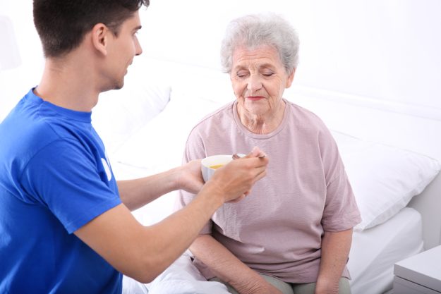 Young male volunteer feeding elderly woman in light room How Nutritional Needs Change As You Age & How Assisted Living Care In University Park, TX Can Help