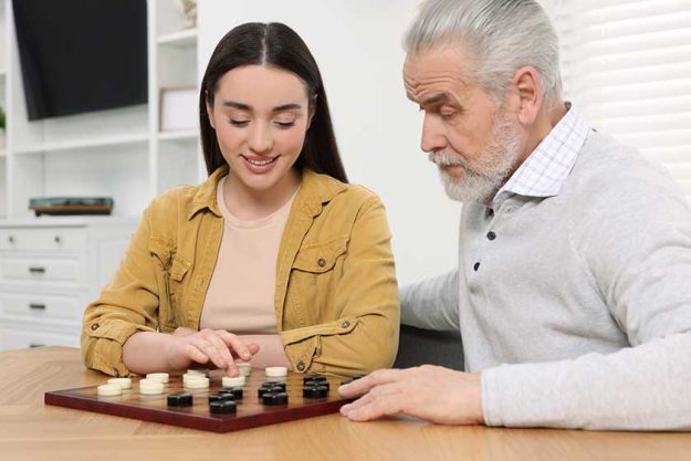 Playing checkers. Senior man learning young woman at table in room